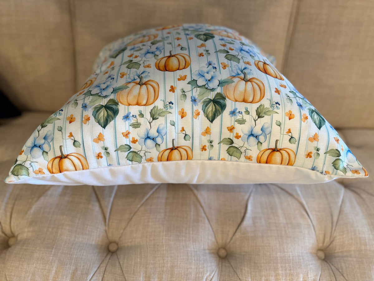 A decorative throw pillow with a fall harvest pattern featuring orange pumpkins, blue flowers, and green leaves on a white striped background, styled on a beige tufted sofa.
