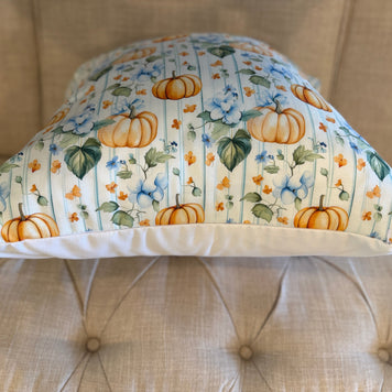 A decorative throw pillow with a fall harvest pattern featuring orange pumpkins, blue flowers, and green leaves on a white striped background, styled on a beige tufted sofa.