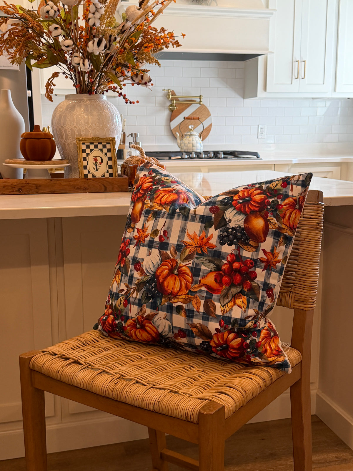 A decorative throw pillow with an autumn harvest print featuring pumpkins, gourds, and berries over a navy and beige gingham pattern, resting on a woven wood chair in a bright kitchen setting.