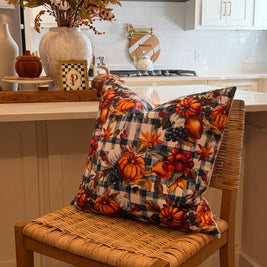 A decorative throw pillow with an autumn harvest print featuring pumpkins, gourds, and berries over a navy and beige gingham pattern, resting on a woven wood chair in a bright kitchen setting.